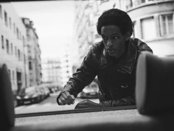 Monochrome portrait of a black man wearing a leather jacket leaning into a car on a city street