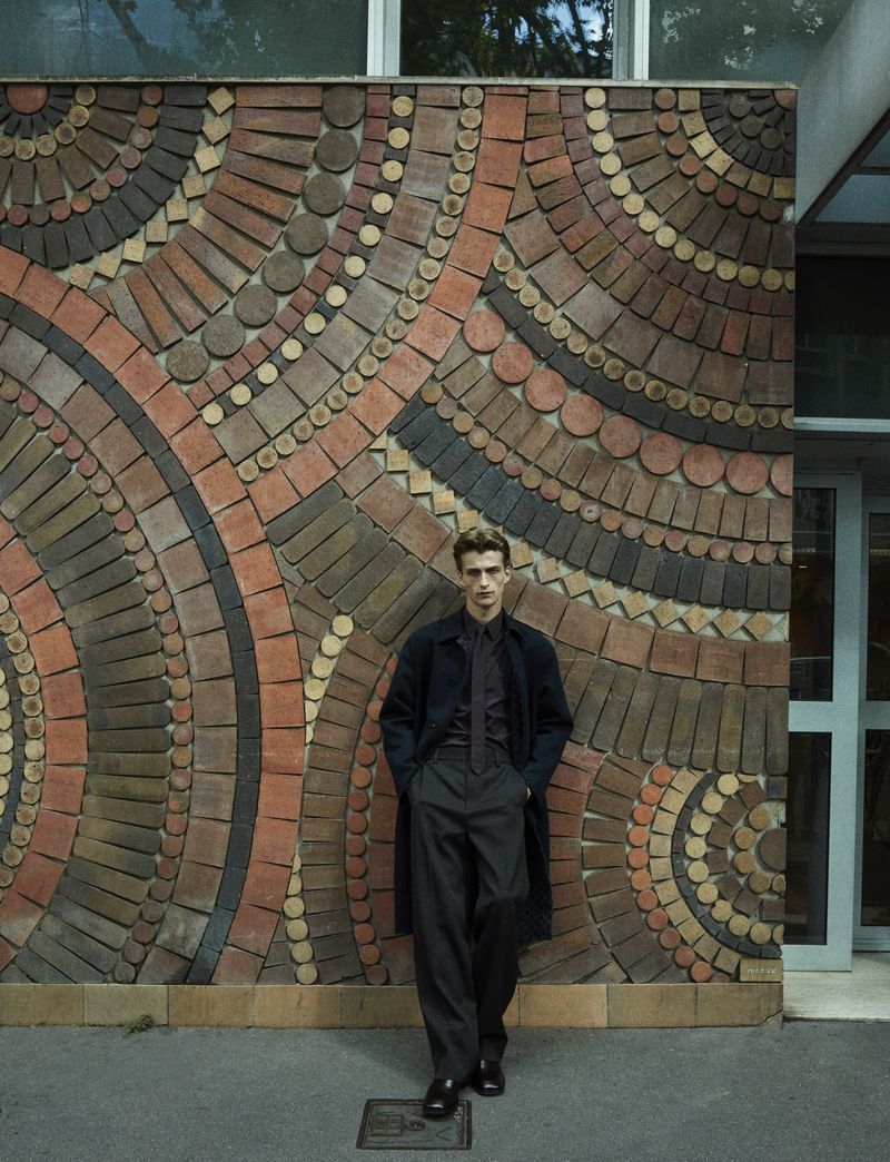 Adult male model stands against a brown and tan patterned wall wearing a dark colored coat, shirt and pants in an urban environment.