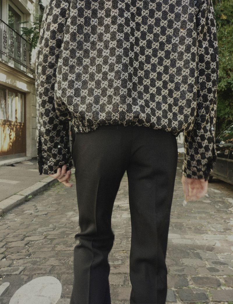 From the back, a person on a cobblestone street wears black pants and a jacket with a dark interlocking logo pattern.