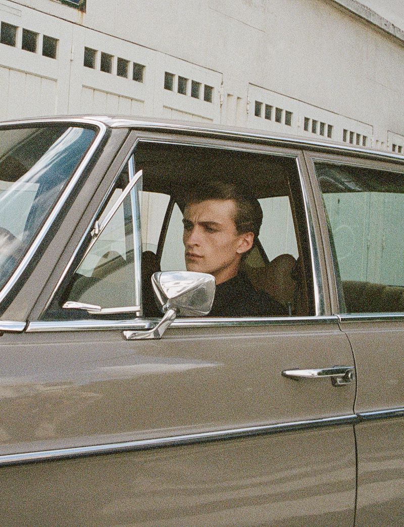 Young man driving vintage tan car with white building and windows in background, cinematic outdoor daytime lifestyle automotive scene.