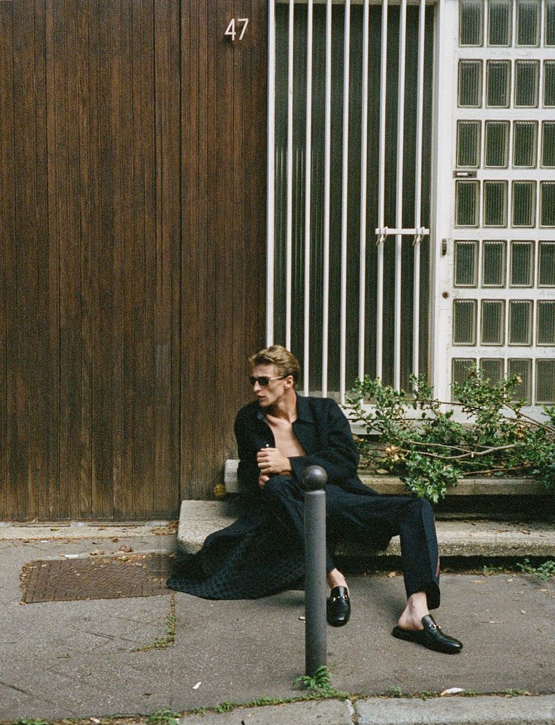 A blonde male fashion model in sunglasses and a dark coat sits on concrete steps outside a building wearing stylish black leather mules.
