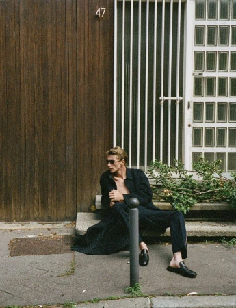 A blonde male fashion model in sunglasses and a dark coat sits on concrete steps outside a building wearing stylish black leather mules.