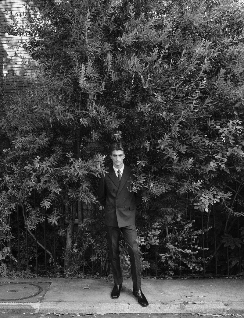 Man standing in a dark suit before dense green foliage on a paved sidewalk for Dapper Dan Magazine.