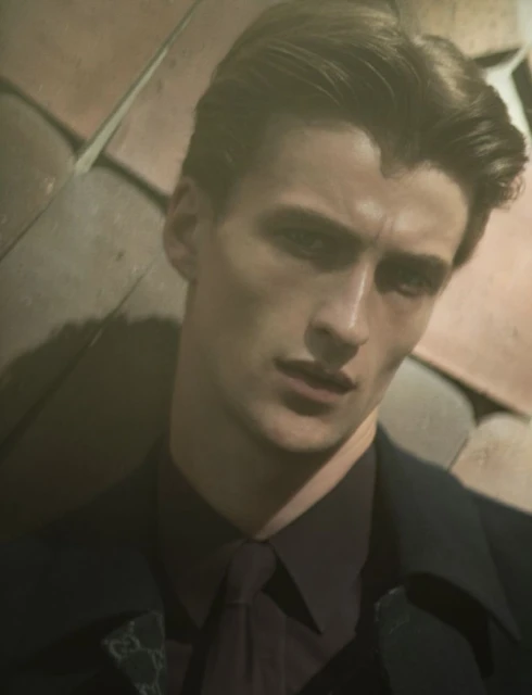 Headshot of a dark-haired adult male wearing a collared shirt and suit jacket, posing indoors against a textured background in soft lighting.