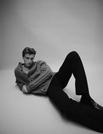 Black and white portrait of a young adult male laying on the ground wearing a cable knit cardigan, trousers, socks, and loafers in a studio.