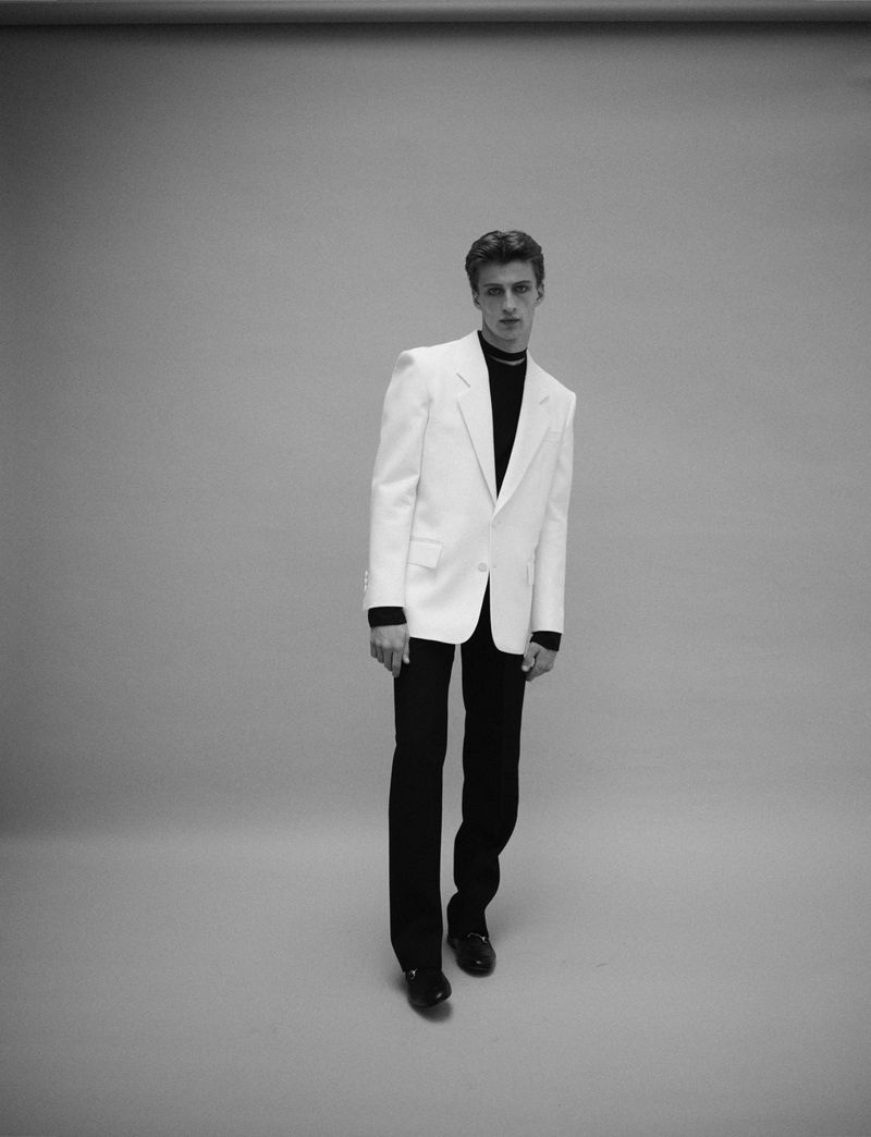 Model wearing white suit jacket and dark trousers standing against a plain grey studio backdrop.