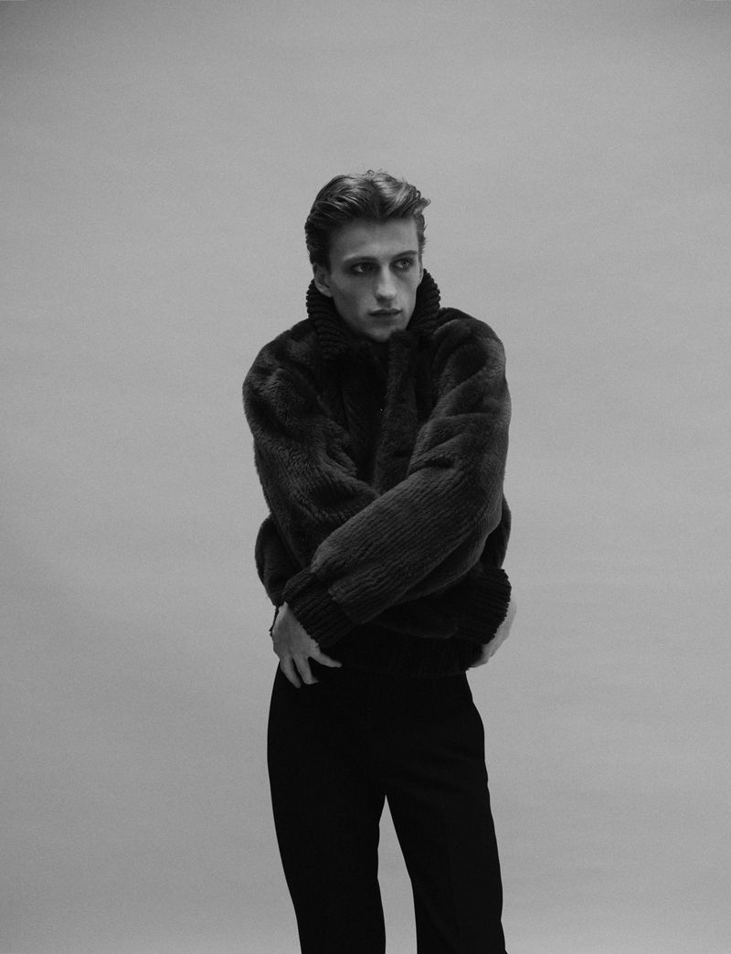Monochrome portrait of a young male fashion model wearing a plush corduroy jacket and dark pants in a photo studio setting.