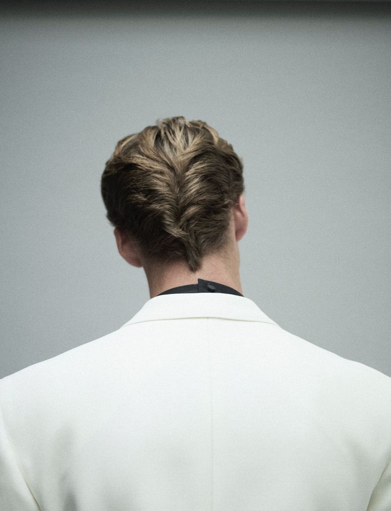 Rear view of a man with styled hair wearing a white jacket against a grey background.