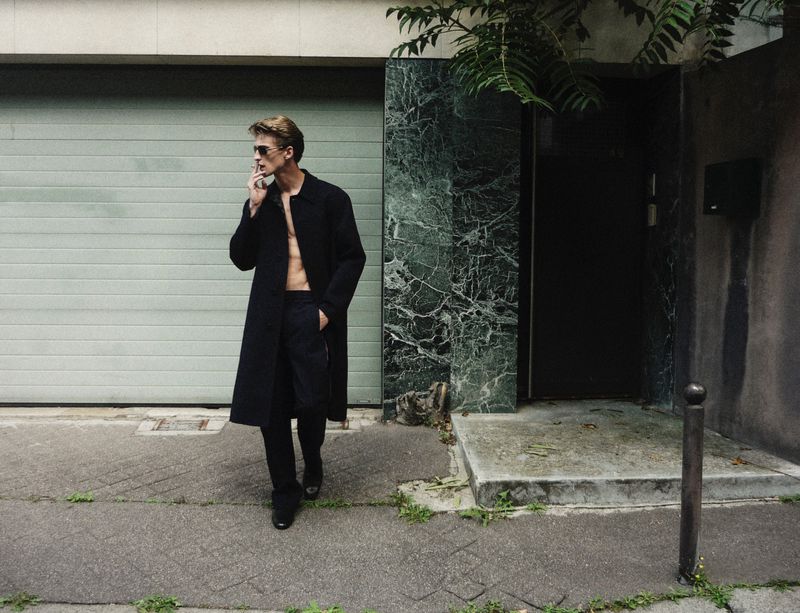A blonde adult male wearing a black overcoat walks on a sidewalk in an urban setting and smokes a cigarette next to a building.