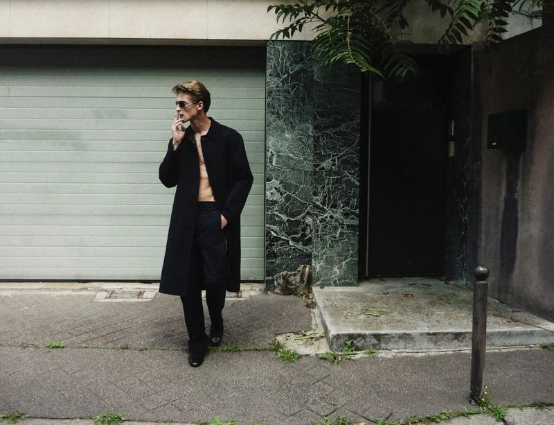A blonde adult male wearing a black overcoat walks on a sidewalk in an urban setting and smokes a cigarette next to a building.