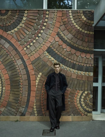 Model wearing dark coat and trousers standing against a patterned brick mosaic wall for Dapper Dan Magazine.