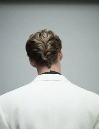 Rear view of a man with styled hair wearing a white jacket against a grey background.