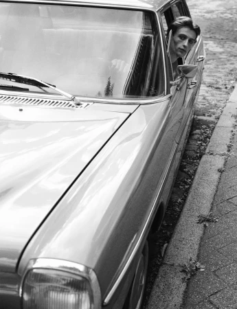 A man looking out the open window of a parked vintage car on a cobblestone street, photographed in black and white.