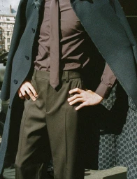 Model standing outdoors wearing a dark coat, brown shirt, matching tie, and trousers with hands on hips