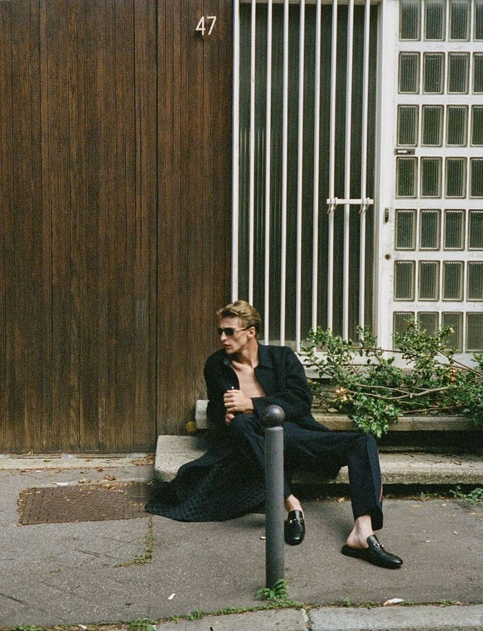 Man wearing dark sunglasses and black coat seated on stone steps outside a wooden doorway.
