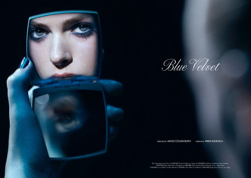 Beauty shot of an adult woman with blue makeup reflected in a hand mirror against a dark backdrop, studio lighting, and makeup details.
