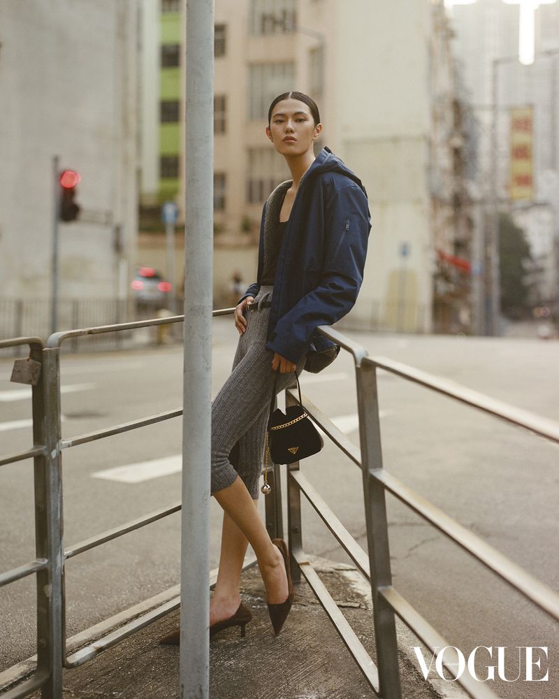 Asian woman in blue jacket poses on city street leaning against railing with Prada purse, gray leggings, brown high-heeled shoes, traffic light.