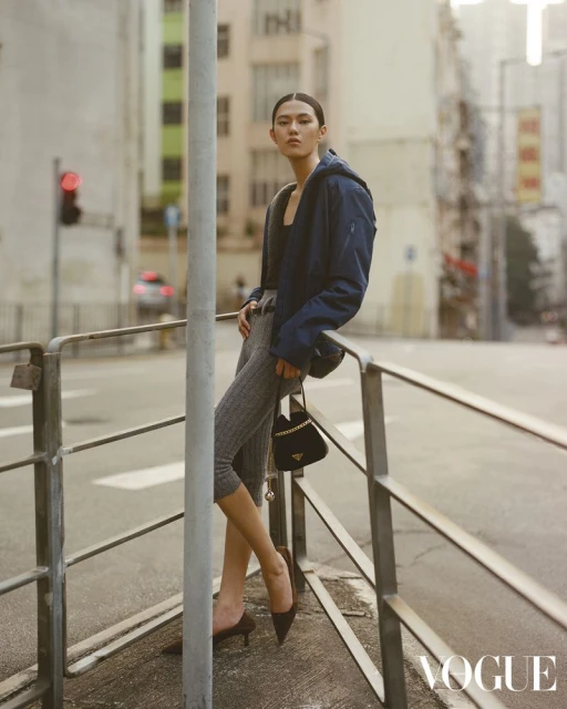 Asian woman in blue jacket poses on city street leaning against railing with Prada purse, gray leggings, brown high-heeled shoes, traffic light.