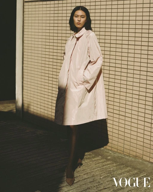 A stylish woman in a pink coat poses against a textured wall, complemented by natural light and shadows, in an outdoor fashion editorial.