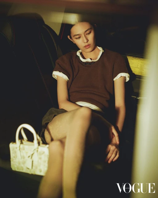 Woman poses in brown top, white trim; handbag sits near. Interior car setting, soft light. Vogue logo. Fashion, beauty.