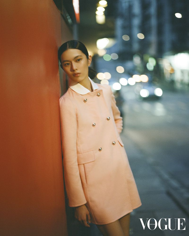 A fashionable Asian woman in a peach coat leans against an orange wall at night, with city lights blurred in the background.
