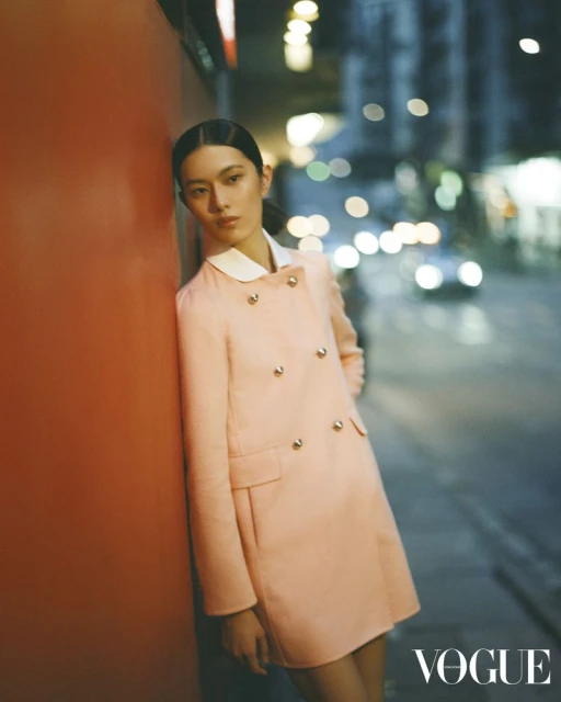 A fashionable Asian woman in a peach coat leans against an orange wall at night, with city lights blurred in the background.