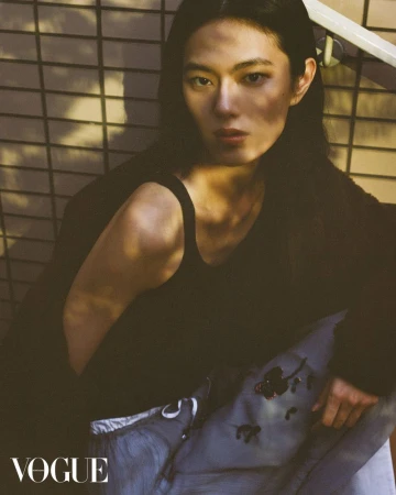 Indoor portrait of an adult male with dark hair, wearing a tank top and sweatpants, posing against a wall with a grid pattern.