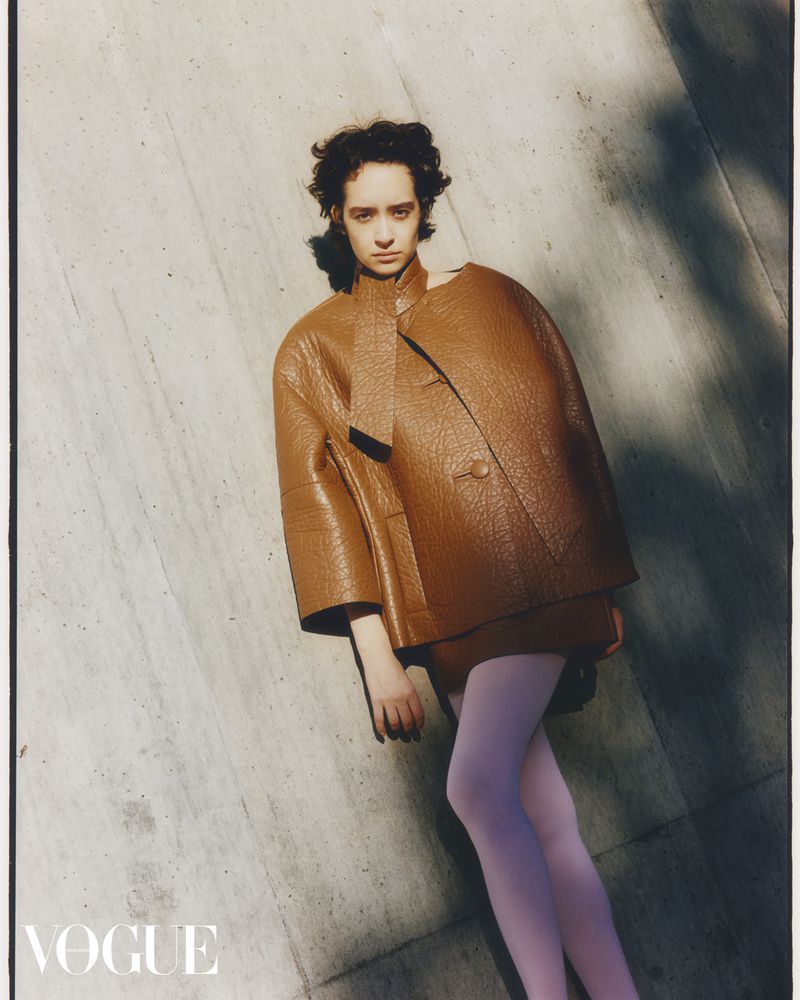 Fashion editorial shot of a woman with short brown hair in a brown leather jacket and pink tights, posing in front of a stone wall.
