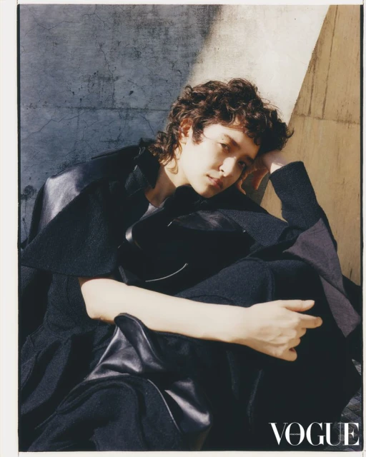Portrait of an adult with curly hair wearing a black coat and pants sitting against a concrete wall, soft lighting and Vogue logo.