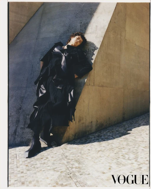 Fashion shot featuring a female model in a black outfit leaning against a concrete wall on a cobblestone surface in an outdoor setting.