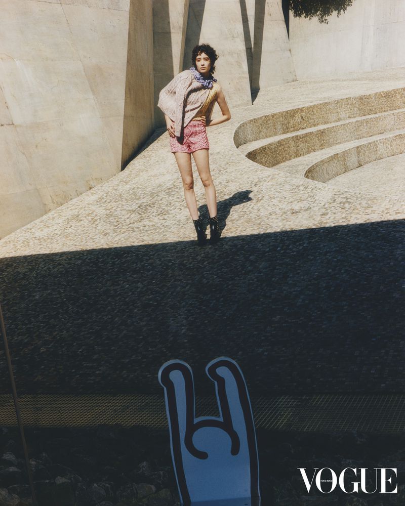 Fashion shot of a woman in patterned shorts and jacket posing on a concrete structure with "Vogue" logo and a blue graphic in the foreground.