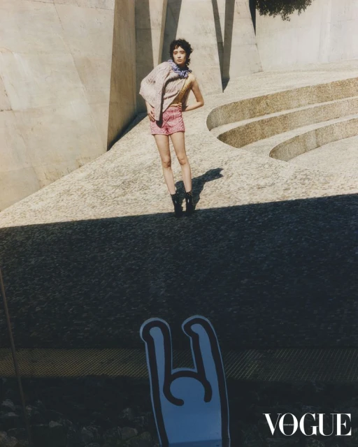 Fashion shot of a woman in patterned shorts and jacket posing on a concrete structure with "Vogue" logo and a blue graphic in the foreground.