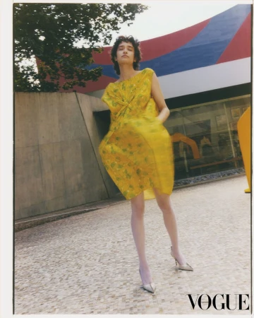 Model wearing a vibrant yellow patterned dress standing outdoors on paved ground for a fashion editorial.