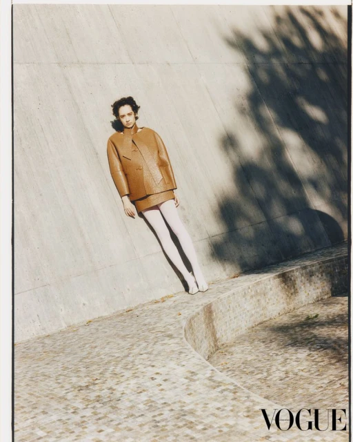 A woman in a tan jacket and skirt poses in a fashion shot against a concrete wall with tree shadows and the Vogue logo in the lower right.