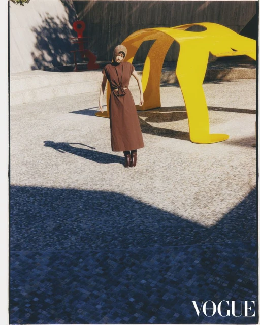 Fashion editorial shot of an adult woman in a brown dress and matching head covering standing in front of yellow abstract outdoor sculpture.