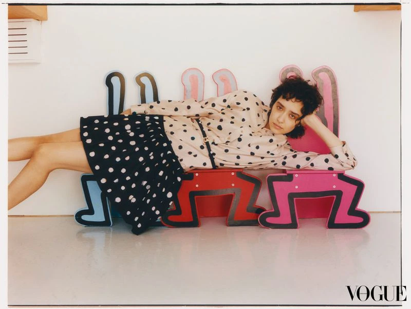A person in a polka dot skirt and blouse poses while reclining on a colorful furniture piece in a bright, minimalist room setting.
