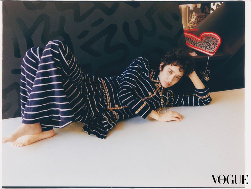 A woman lies on a white surface, wearing a striped blue and white suit with jewelry, in front of a black patterned backdrop.