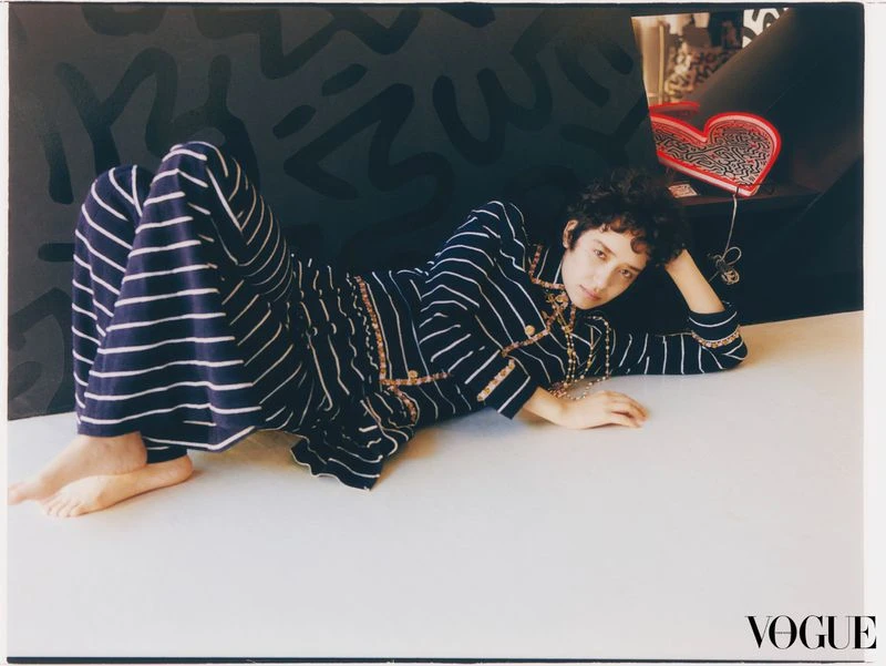 A woman lies on a white surface, wearing a striped blue and white suit with jewelry, in front of a black patterned backdrop.