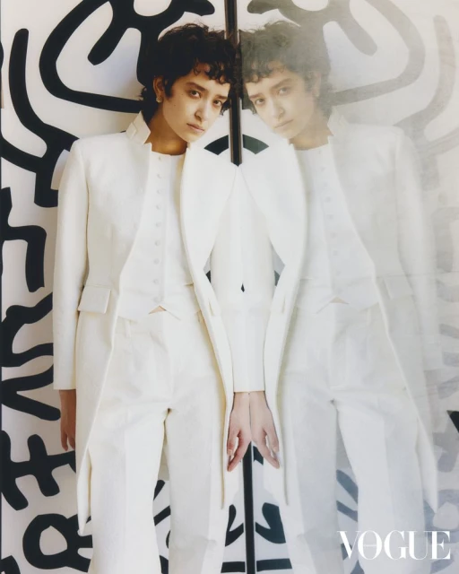 Portrait of a person in a white suit next to a mirror and a patterned wall, gazing at the camera in a Vogue fashion editorial.