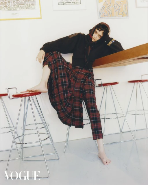 Fashion editorial shot of a slender person with dark hair wearing a hoodie and plaid skirt, sitting on a red stool in a bright room.