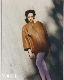 Model in a brown textured coat and pink tights posing against a concrete wall for a Vogue Hong Kong fashion editorial.