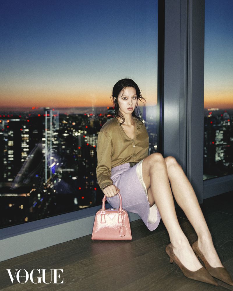 Fashion shot of a woman in a tan shirt and skirt, holding a pink purse, sitting by a window overlooking a cityscape at dusk.