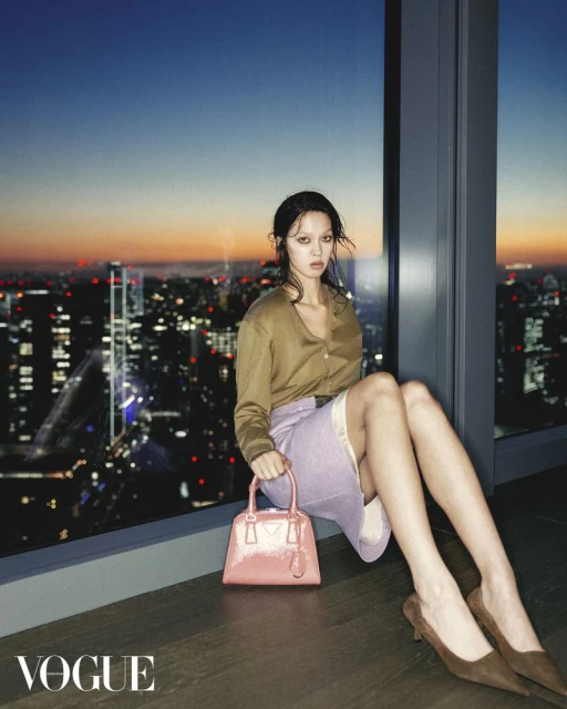 Fashion shot of a woman in a tan shirt and skirt, holding a pink purse, sitting by a window overlooking a cityscape at dusk.