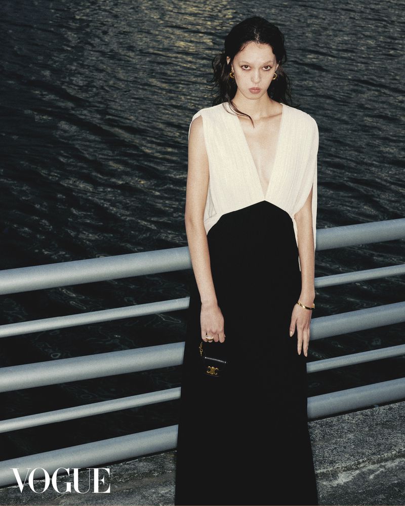 A young woman poses in a sleeveless black and cream dress against a waterfront backdrop, accented by a silver railing and 'Vogue' logo.