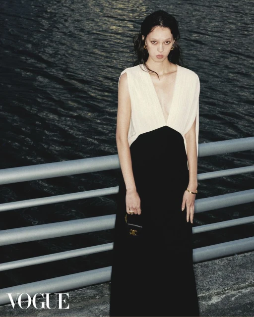 A young woman poses in a sleeveless black and cream dress against a waterfront backdrop, accented by a silver railing and 'Vogue' logo.