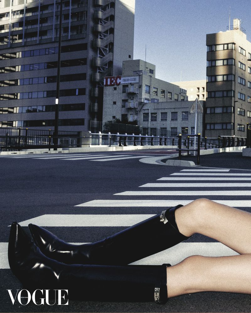 Fashion editorial photo of legs in black leather boots lying on a crosswalk, with tall urban buildings in the background and Vogue logo.