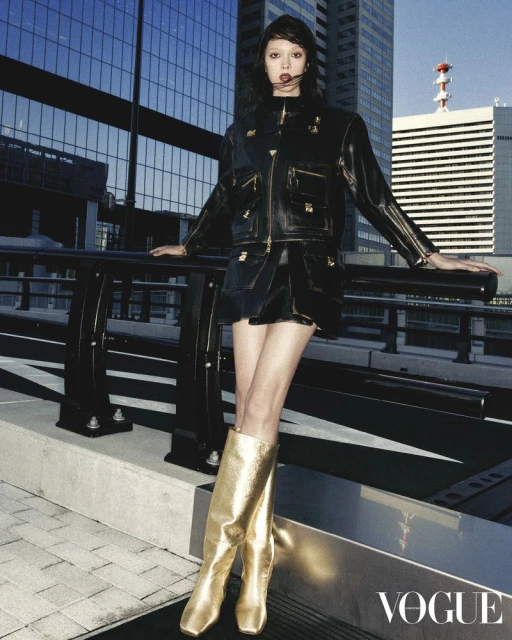 A woman in a black jacket and skirt poses against a city backdrop, wearing gold boots, for a fashion editorial with Vogue logo.