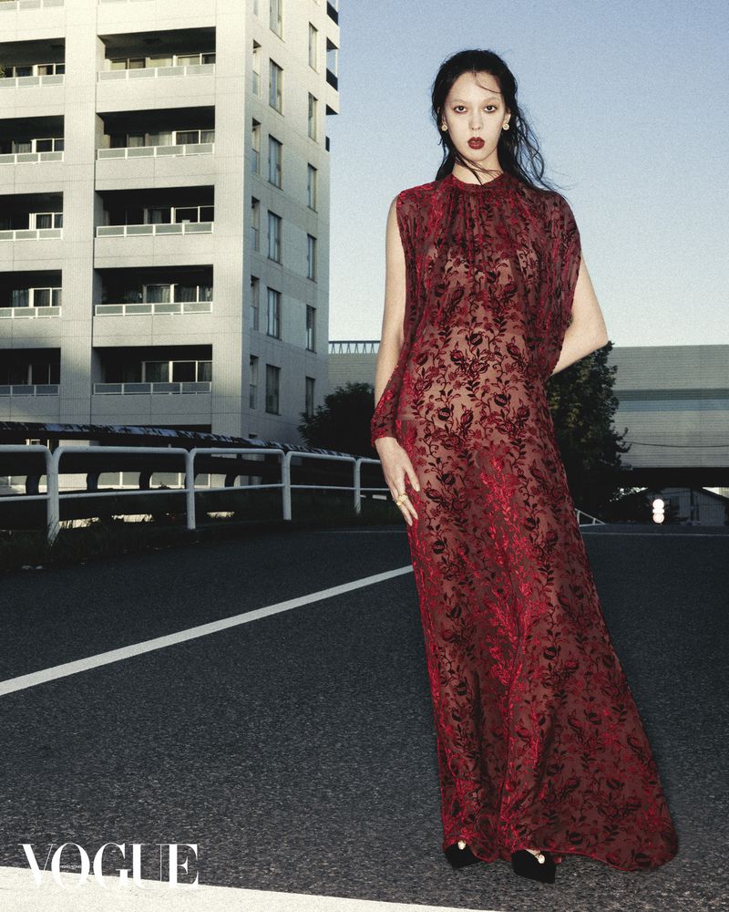A fashionable young woman in a red lace gown poses in front of a modern building on a city street, Vogue logo in corner.