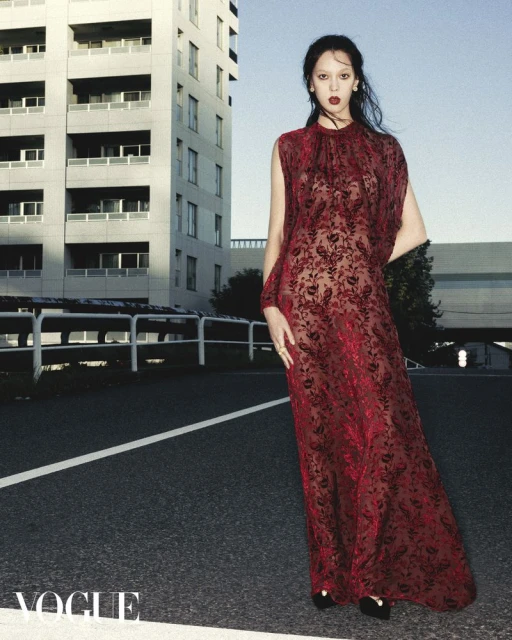 A fashionable young woman in a red lace gown poses in front of a modern building on a city street, Vogue logo in corner.