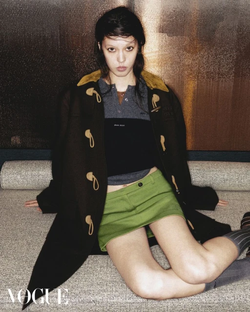 A young woman poses on a textured surface wearing a dark duffel coat, polo shirt, and green skirt in an indoor setting.